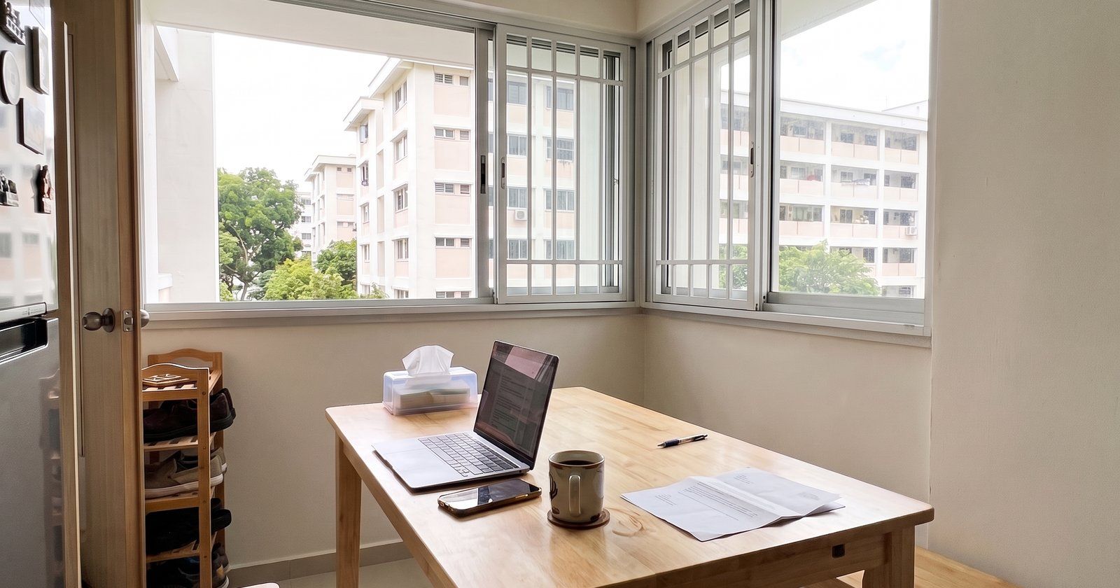 A wooden dining table with a laptop, phone, mug and a printed letter by the corner windows of an HDB flat, with blocks and trees outside